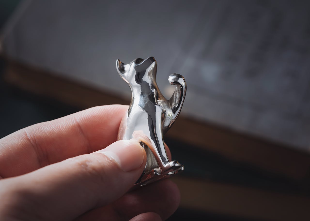Detailed close-up of a shiny silver cat figurine being held, featuring human hand and soft focus background.